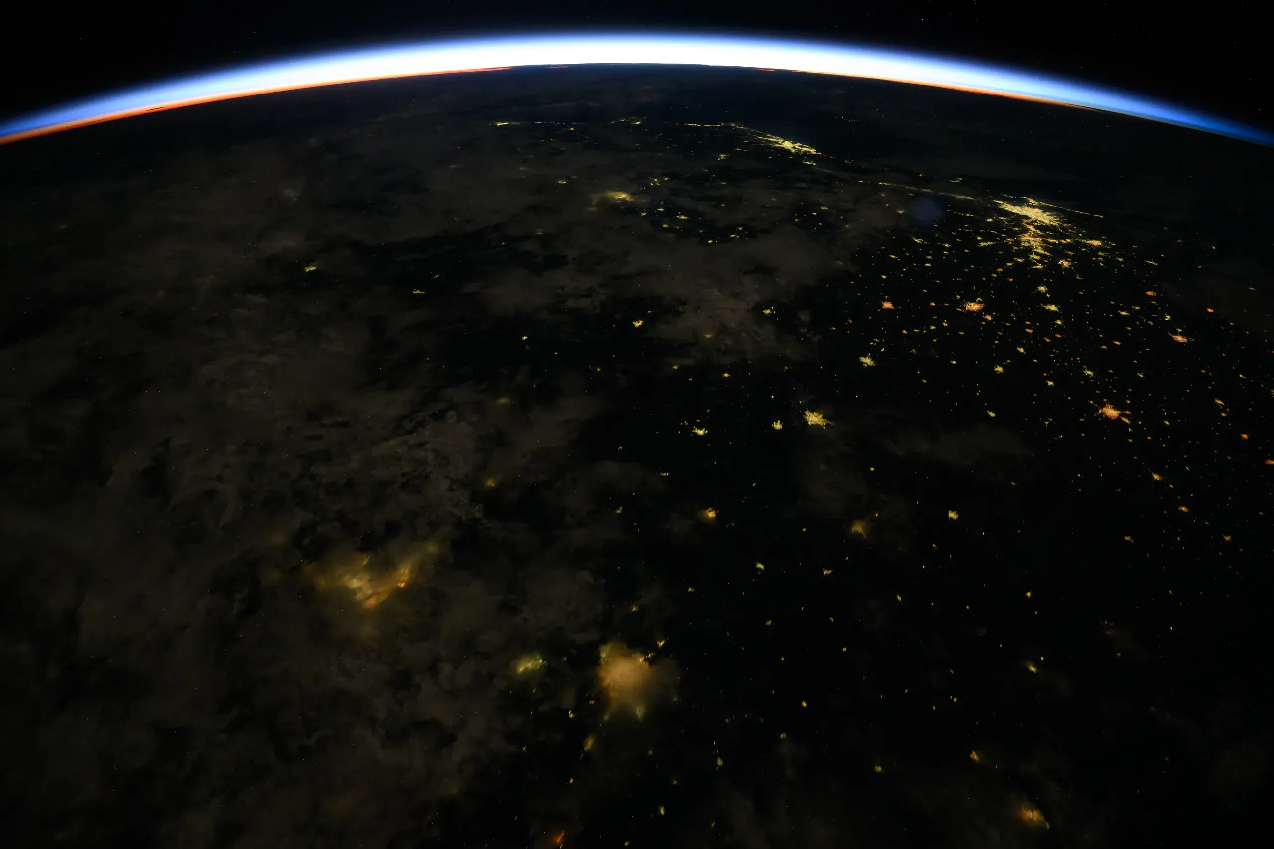 Photograph from orbit at night: the thin blue and orange line of sunrise along the curve of the Earth, with city lights below